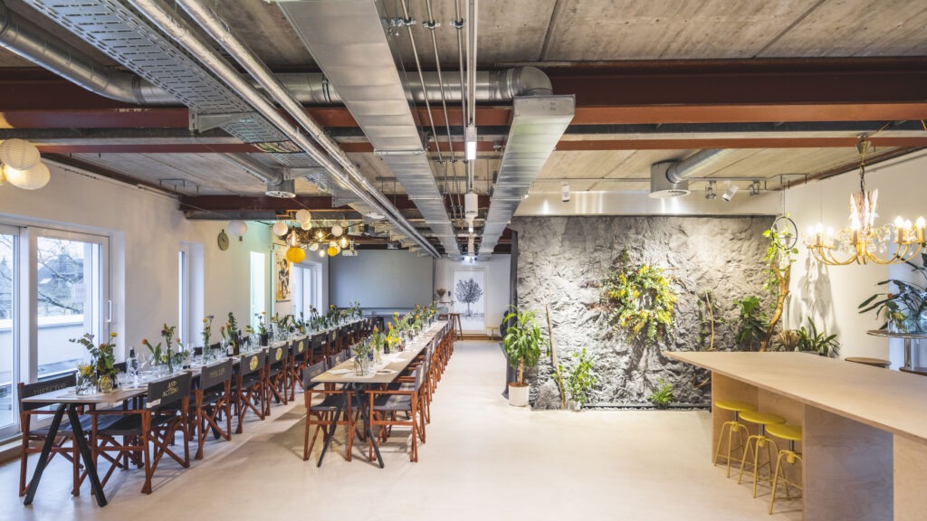 The picture shows the Heinzigartig Loft in the Hotel Luise. You can see two long tables on the left and a large table with several yellow stools on the right. 