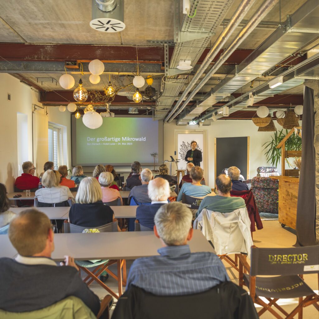 You can see a room full of people sitting in the Heinzigartig Loft in the Hotel Luise, listening to a presentation by Managing Director Ben Förtsch. 